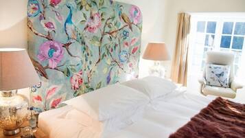 Une chambre à coucher avec une tête de lit à motifs floraux, un lit recouvert de draps blancs, un plaid marron, une table de chevet avec une lampe, une chaise à motifs de papillons et une fenêtre avec des rideaux.