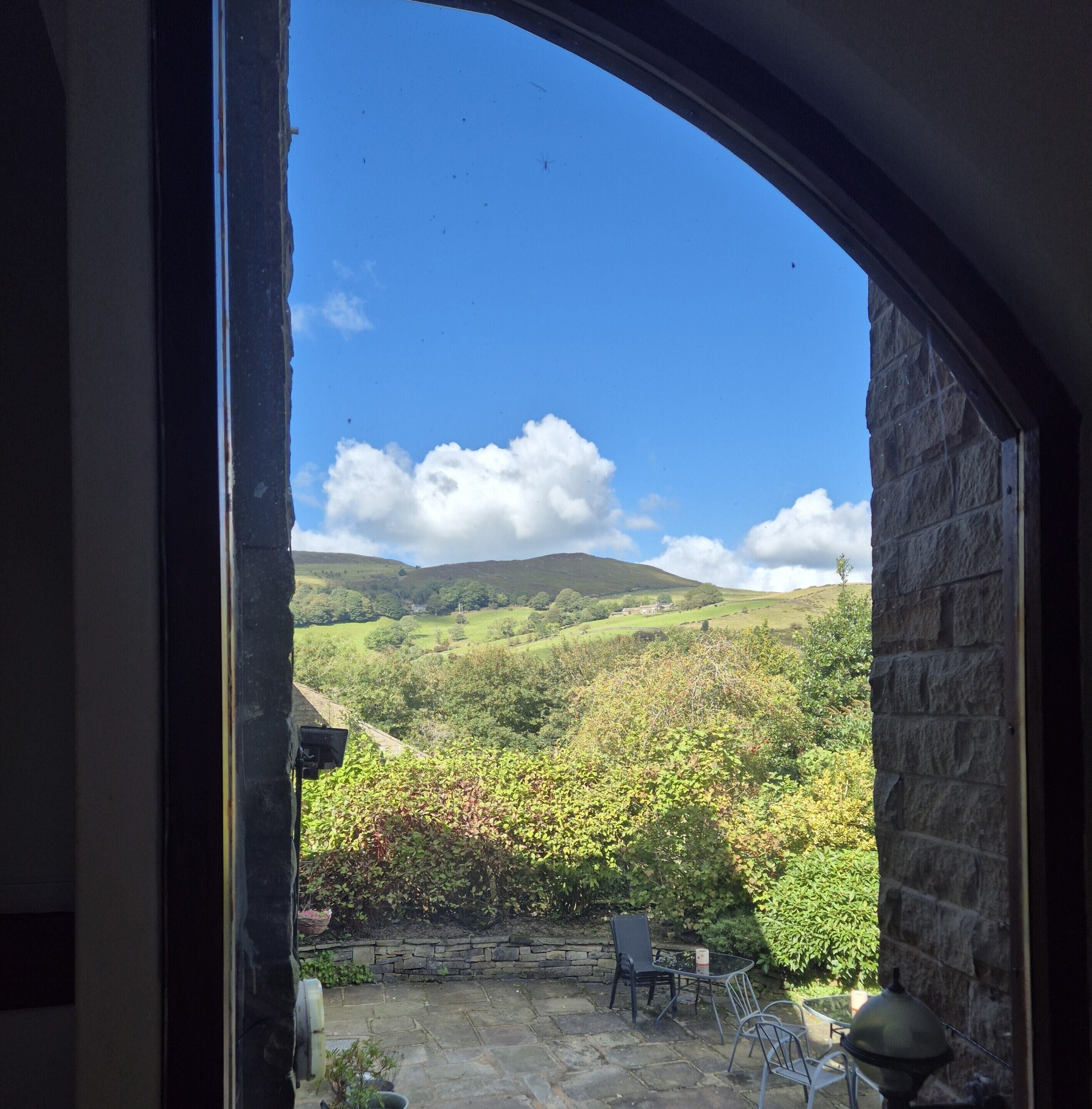 A view through an arched doorway of a stone building with a patio area, stone walls, and a landscape of rolling hills and greenery.