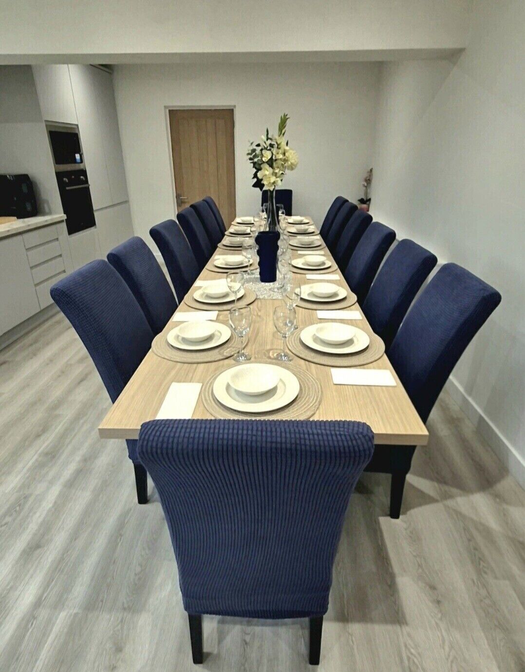 A long dining table set for a meal with blue upholstered chairs, a vase with flowers, and a wooden floor.