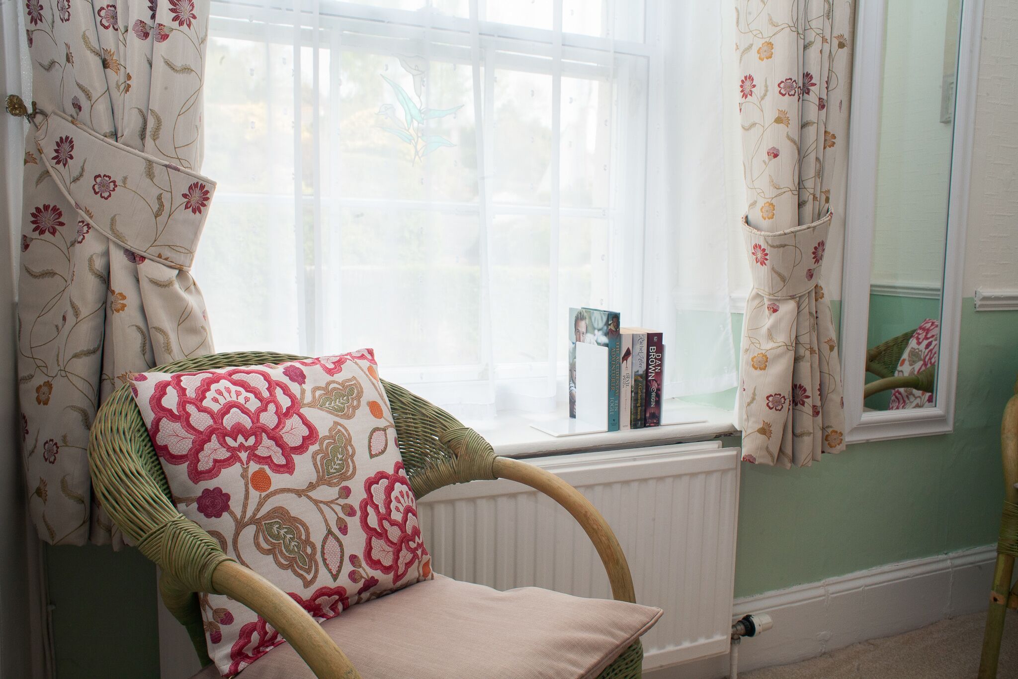 A wicker chair with a floral cushion, a window with curtains, and books on a windowsill.