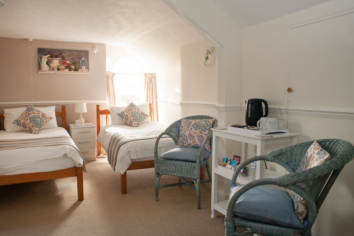 A bedroom with two beds, a wicker chair, a small table with a kettle, and a painting on the wall.