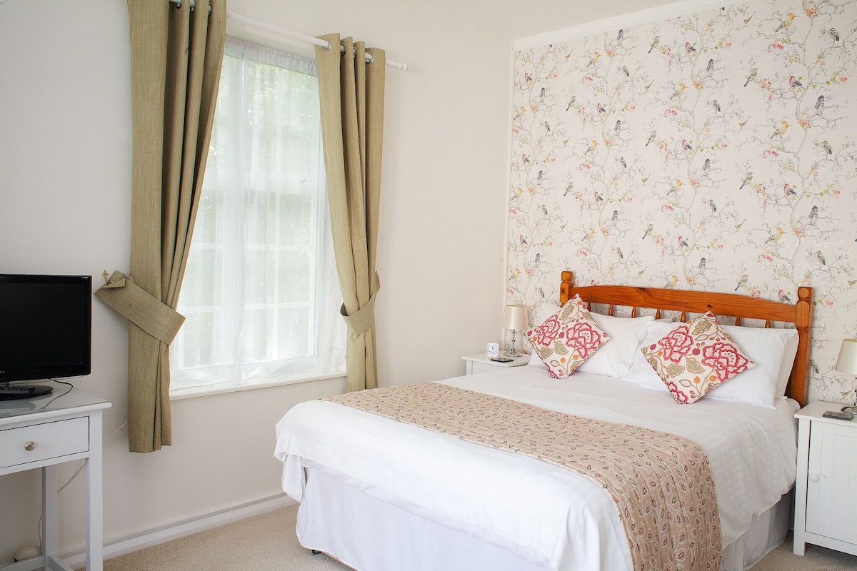 A bedroom with a bed, a bedside table, a TV, and a window with curtains.