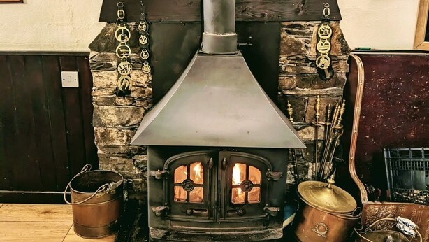 A wood-burning stove with a stone wall backdrop, a metal bucket, and a copper pot.