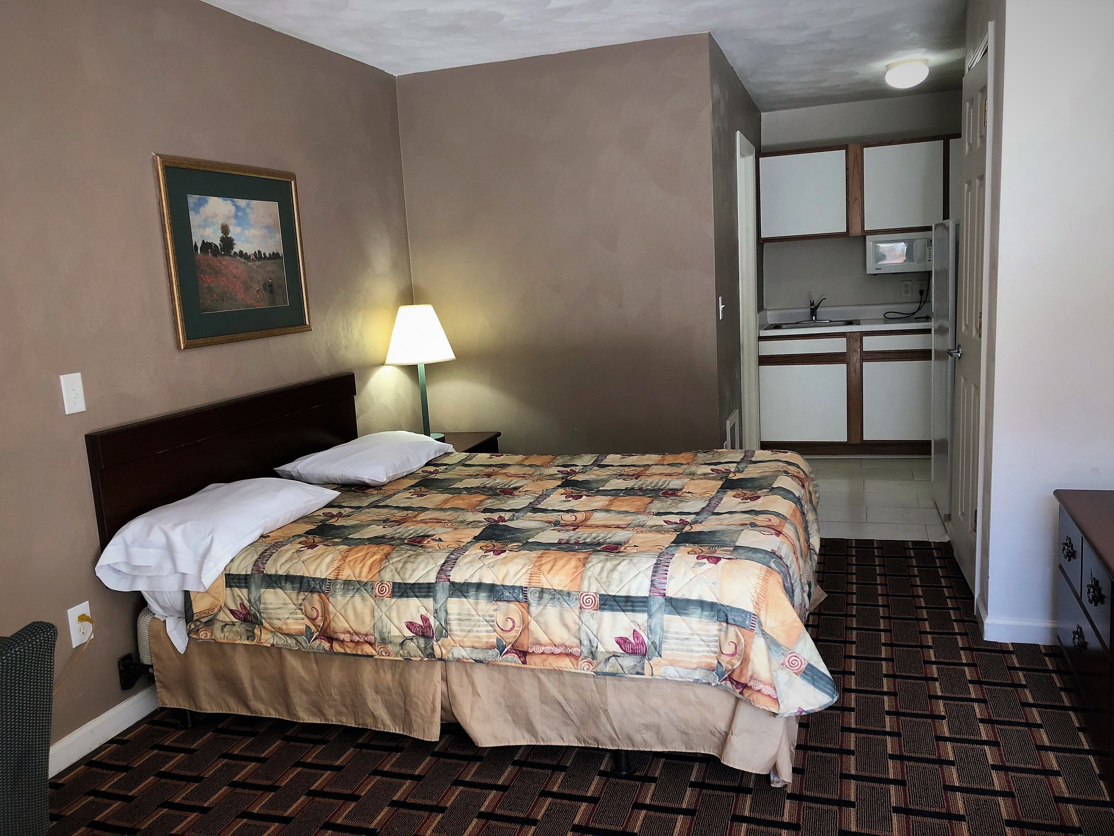 A hotel room with a bed, a nightstand, a lamp, a painting, and a kitchenette.