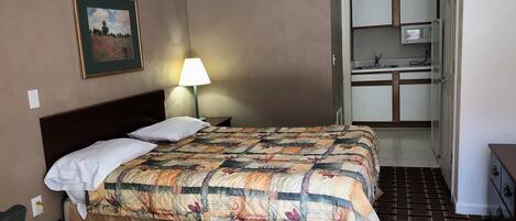 A hotel room with a bed, a nightstand, a lamp, a painting, and a kitchenette.