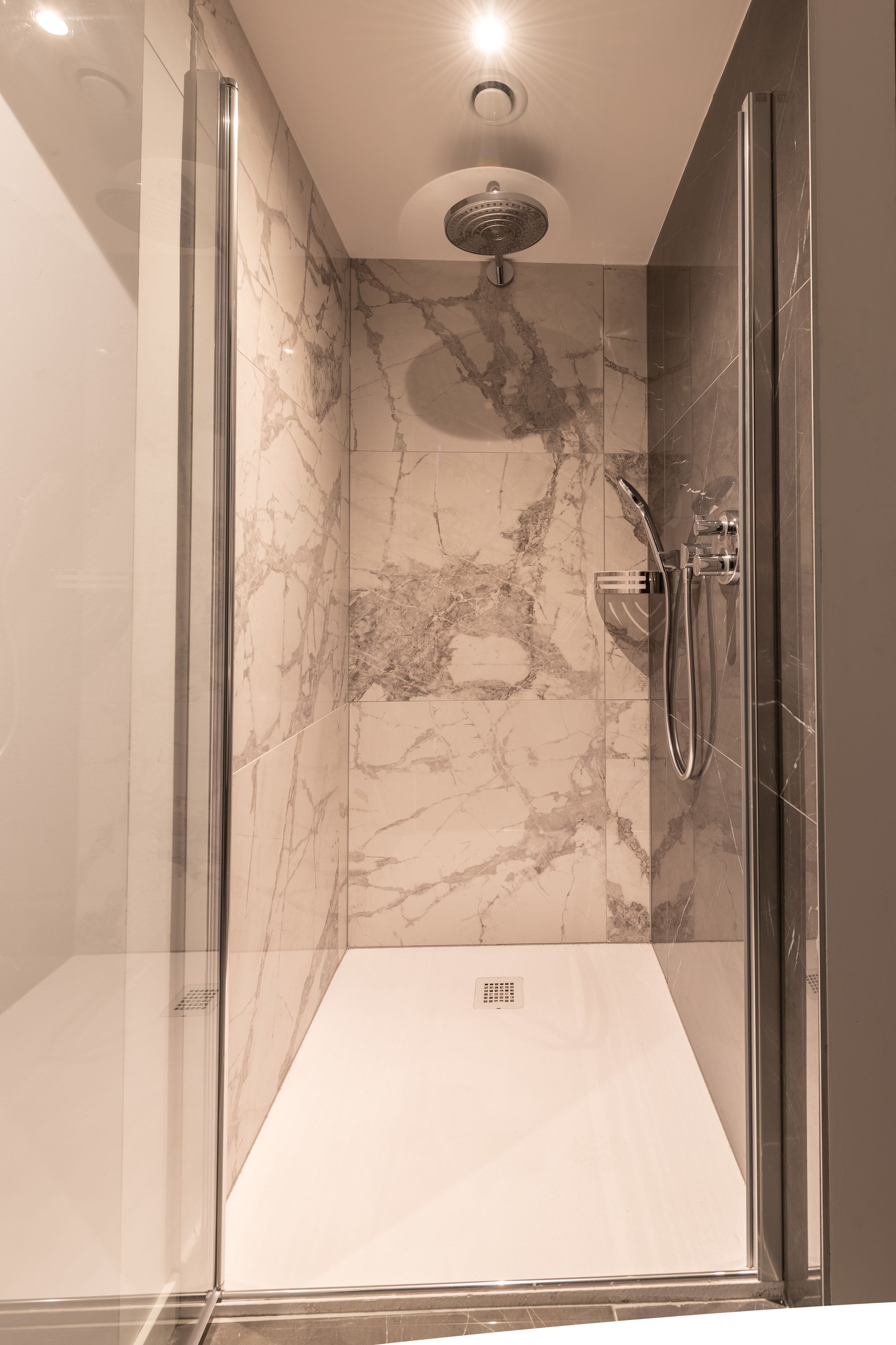 A modern shower with a glass door, marble walls, and a ceiling-mounted showerhead.
