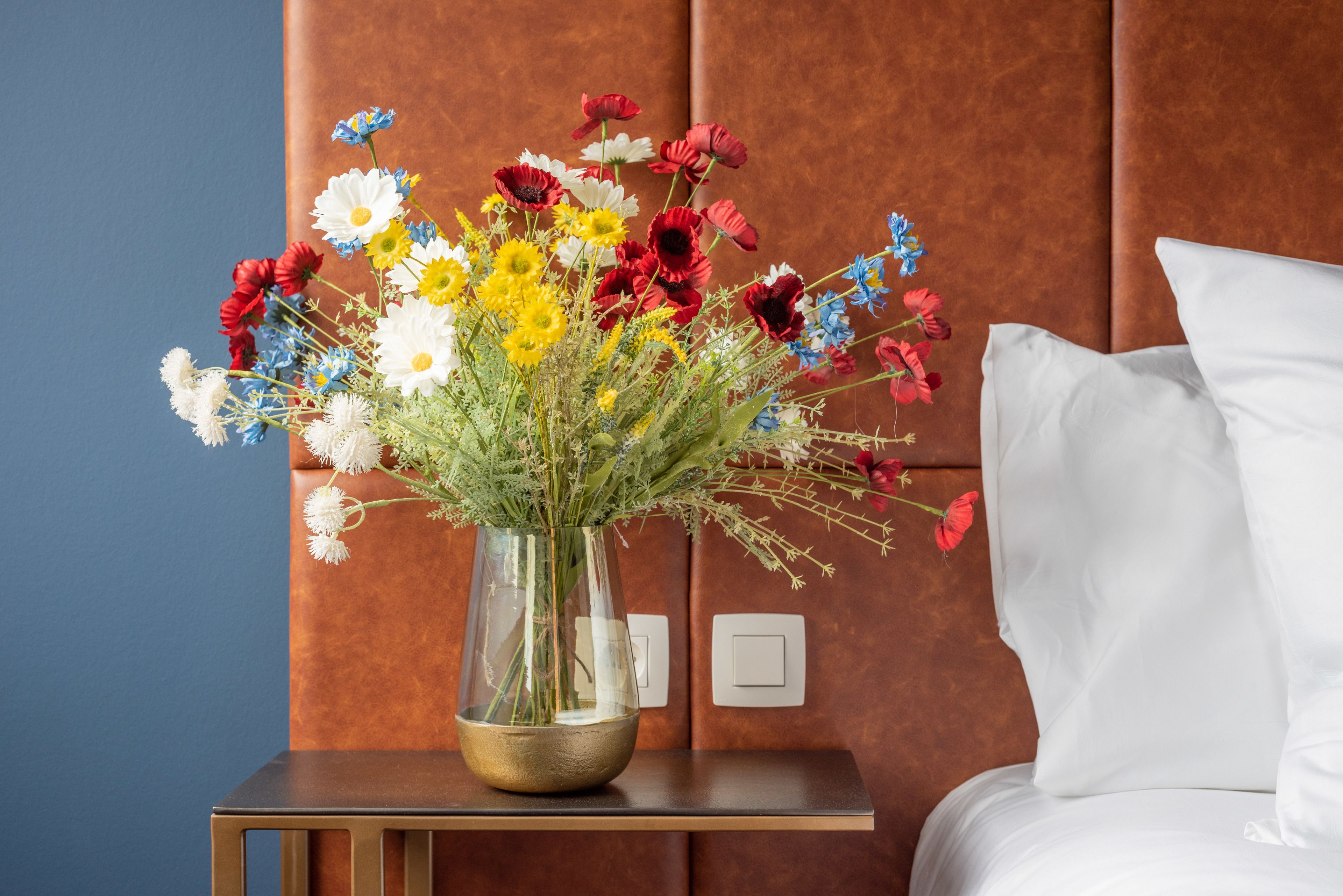 A bed with white linens, a nightstand with a clear vase of flowers, and a brown leather headboard.