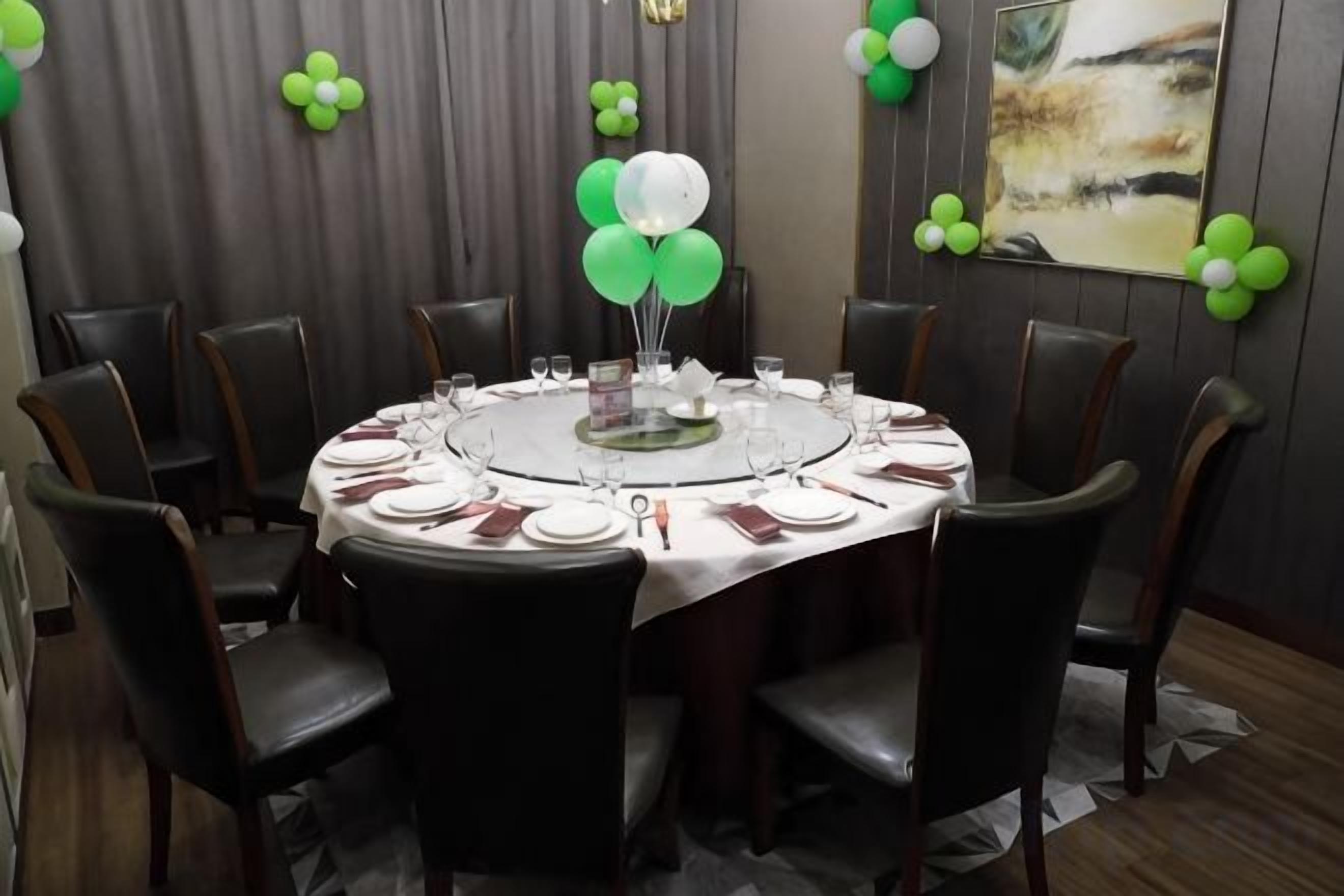 Una mesa de comedor redonda con sillas, dispuesta para una comida, en una habitación con cortina gris y decoración de globos verdes.