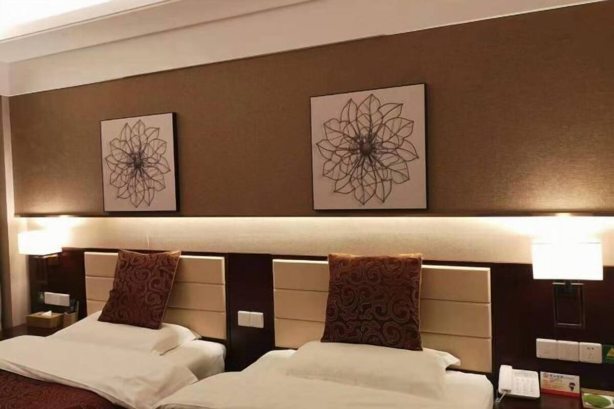 A hotel room with two beds, headboards, wall art, bedside lamps, and a nightstand with a lamp.