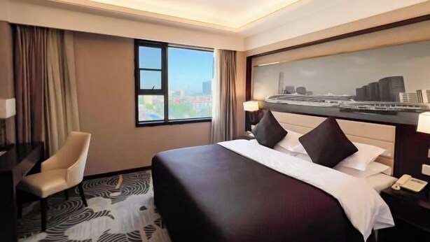 A hotel room with a bed, a desk, a chair, a window with a cityscape view, and a painting on the wall. - Hongluyuan Hotel (Shanghai)