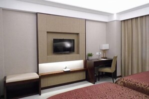 Una camera d'albergo con un letto, una scrivania con una sedia, una TV a parete e una finestra con tende.