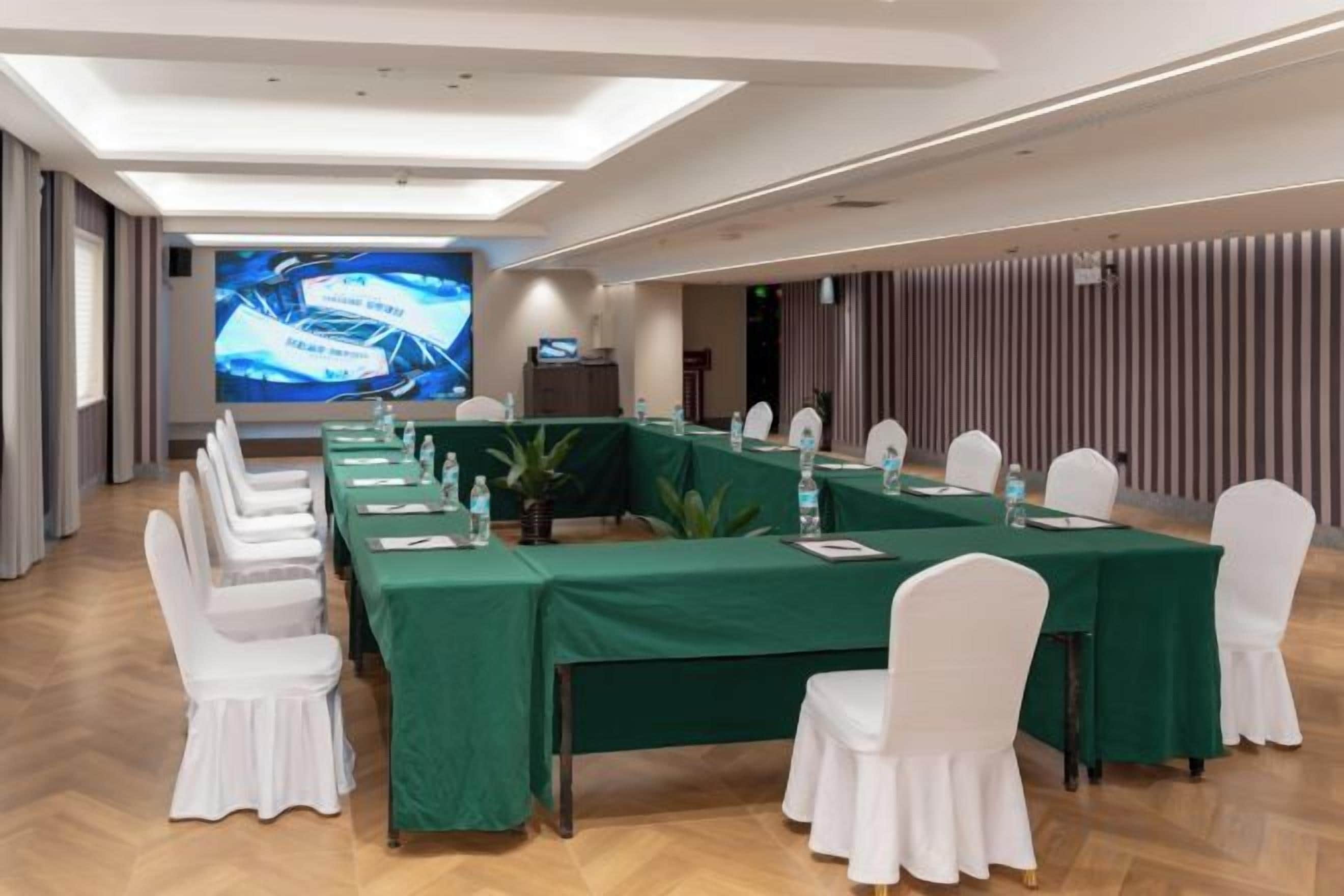 Un salón de conferencias con una mesa larga, sillas blancas y una pantalla de proyección.