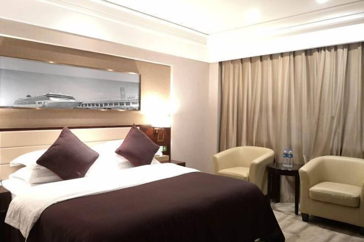 A hotel room with a large bed, two armchairs, a nightstand, a lamp, a painting, and a view of a building.