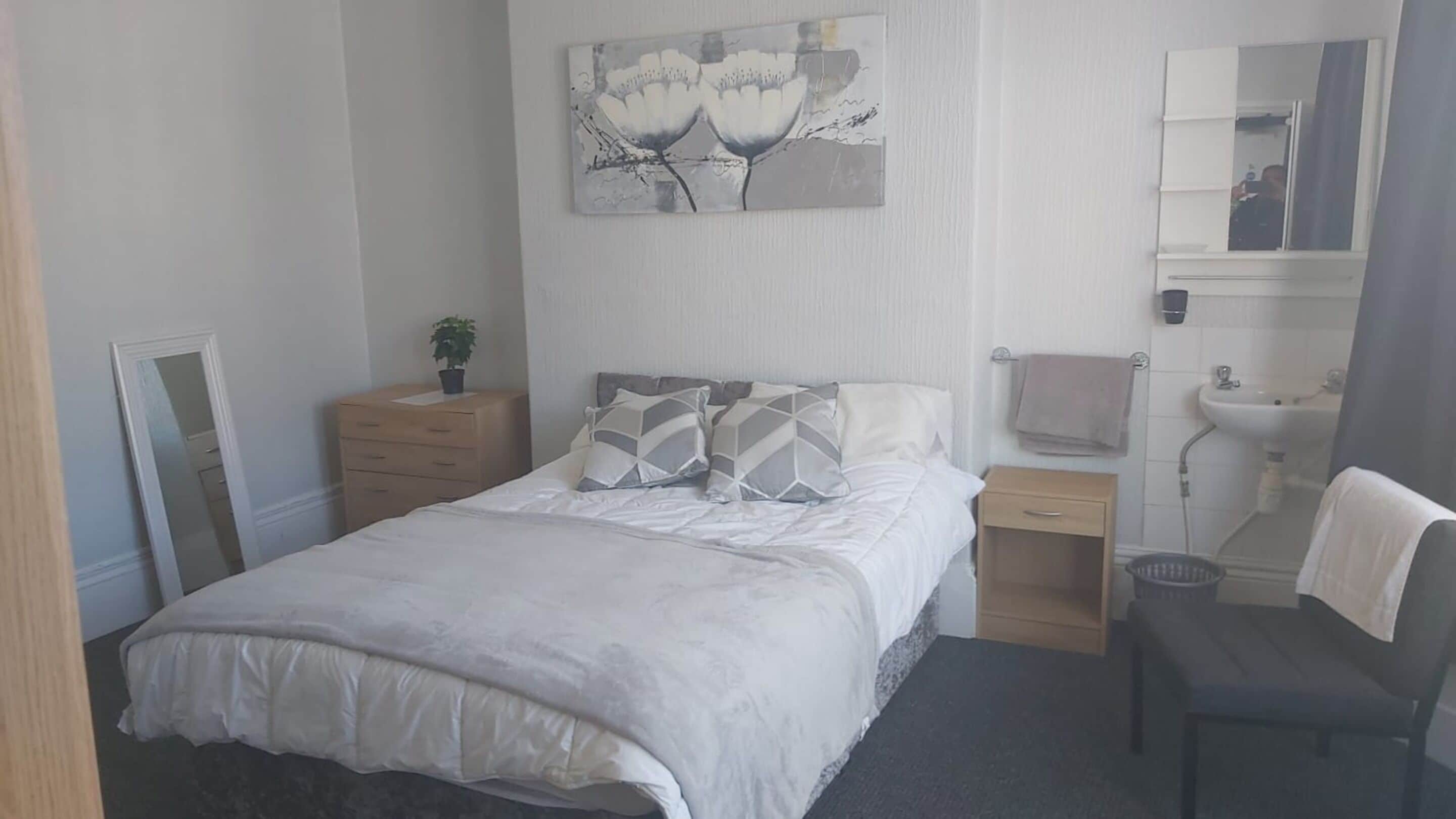 Ein Schlafzimmer mit einem Bett, Nachttischen, einem Spiegel, einem Waschbecken und einem Sessel.