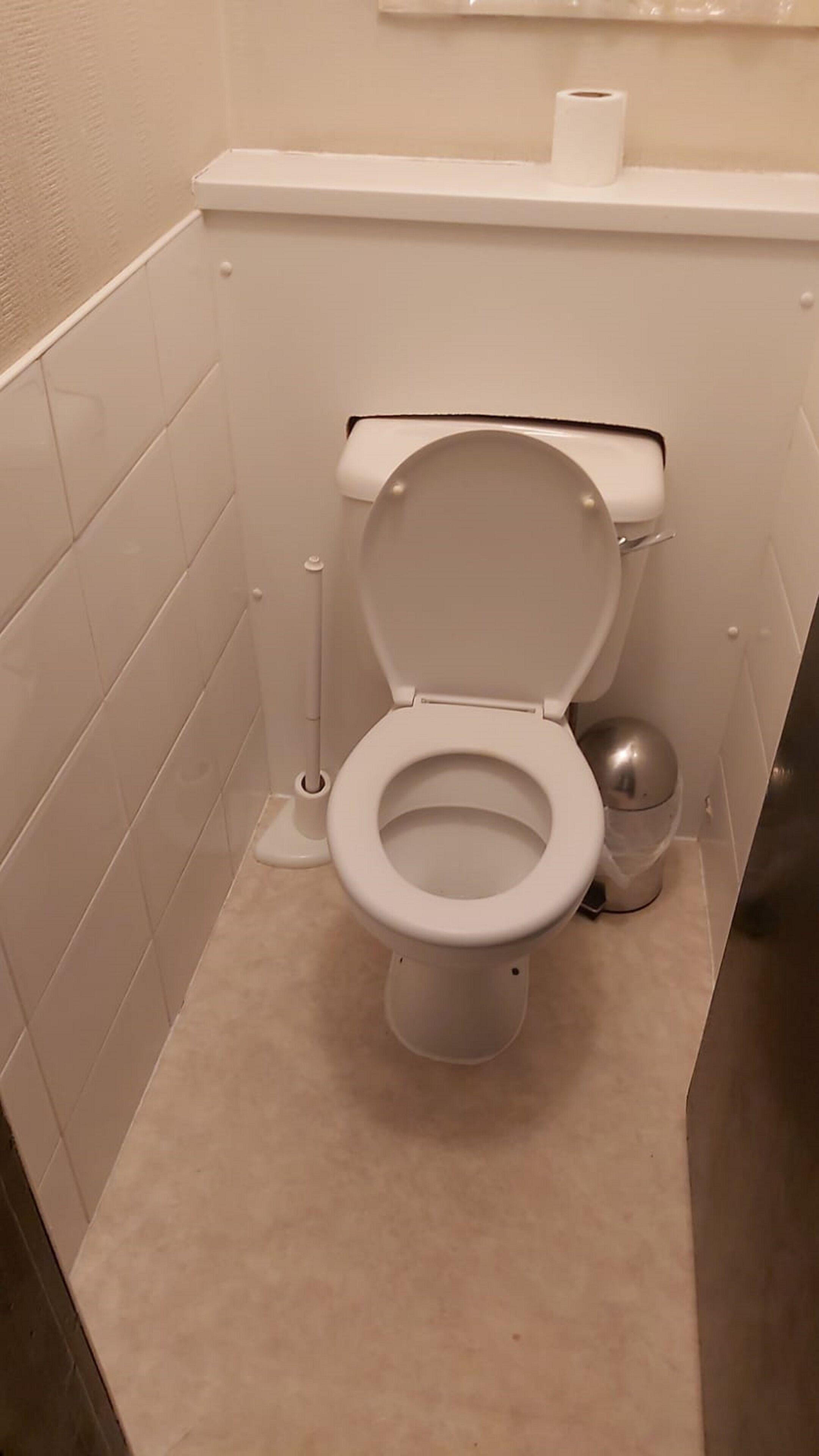 A small, white toilet in a narrow bathroom with beige tiles.