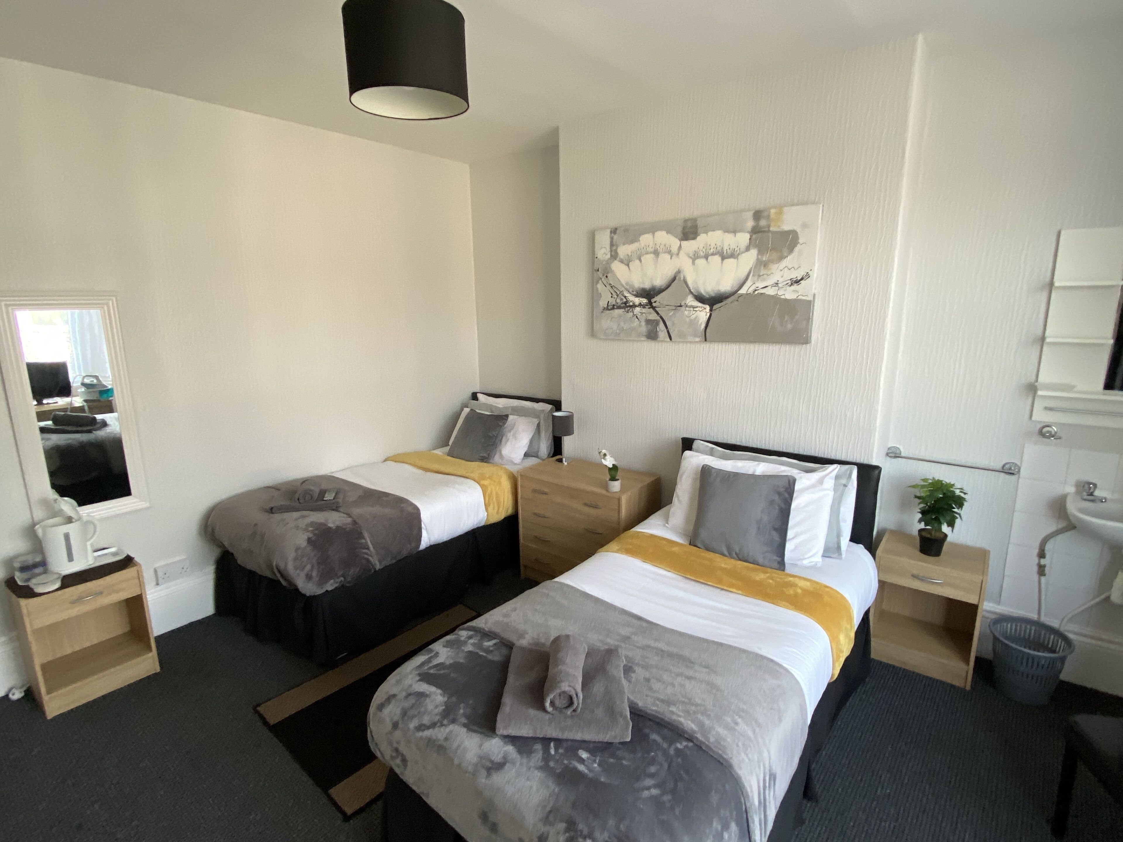 Habitación de hotel con dos camas, una mesita de noche con lámpara, una planta pequeña, un cuadro en la pared y un baño con lavamanos y espejo.