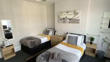 Habitación de hotel con dos camas, una mesita de noche con lámpara, una planta pequeña, un cuadro en la pared y un baño con lavamanos y espejo.