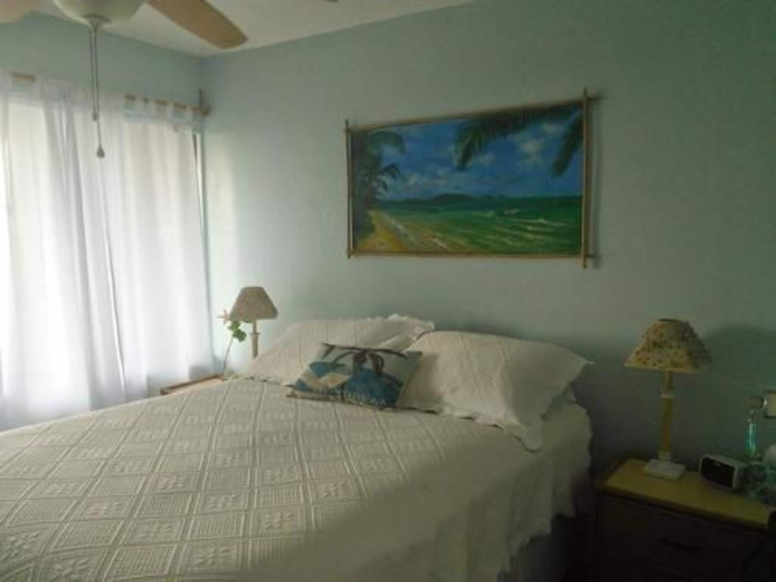Un dormitorio con cama, mesita de noche, lámpara y un cuadro en la pared.
