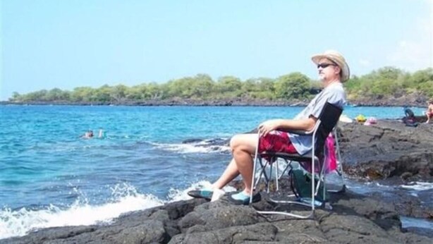 A person sitting on a chair by the water, with a rocky shoreline and trees in the background. - Hale-hoola B & B (Captain Cook)