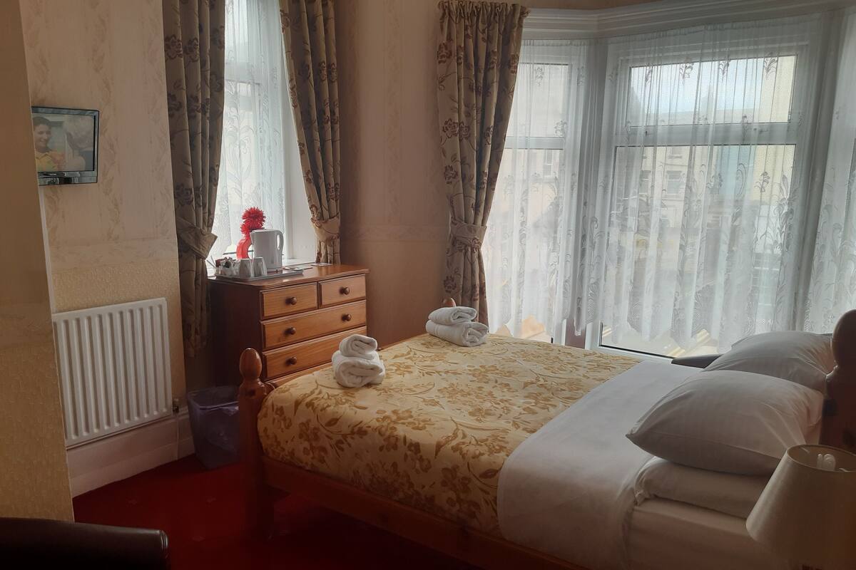 A hotel room with a bed, a wooden dresser, a radiator, and a window with curtains.