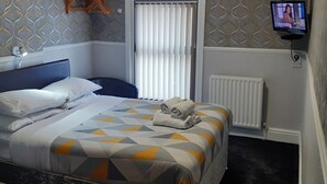 A hotel room with a bed, a television, a window with blinds, a radiator, and a patterned wall. - Wellington Hotel (Blackpool)