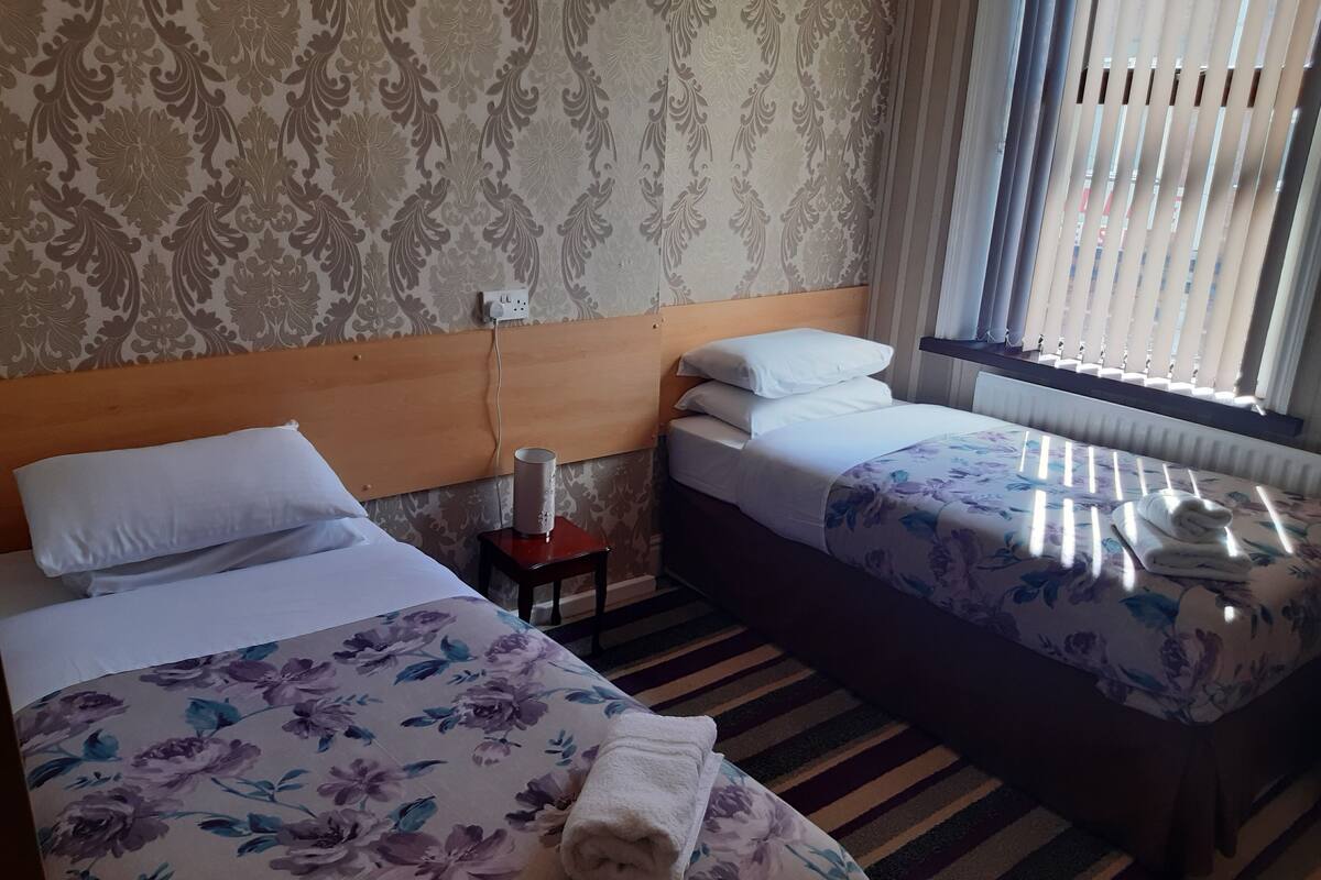 A hotel room with two beds, each with floral bedding and white pillows, a wooden headboard, a small table with a lamp, and a window with blinds.