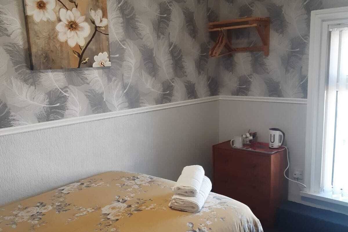 A neatly made bed with a floral bedspread, a wall-mounted artwork of flowers, and a small wooden cabinet with a kettle.