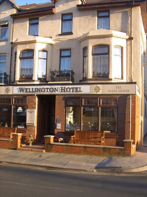 A two-story hotel with a ground floor entrance, a balcony with a wrought iron railing, and a sign indicating the hotel's name and contact number. - Wellington Hotel (Blackpool)