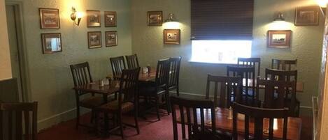 A dining area with wooden tables and chairs, framed pictures on the walls, and a window with blinds.