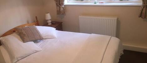 A neatly made bed in a room with a window, a radiator, and a small table with a lamp.