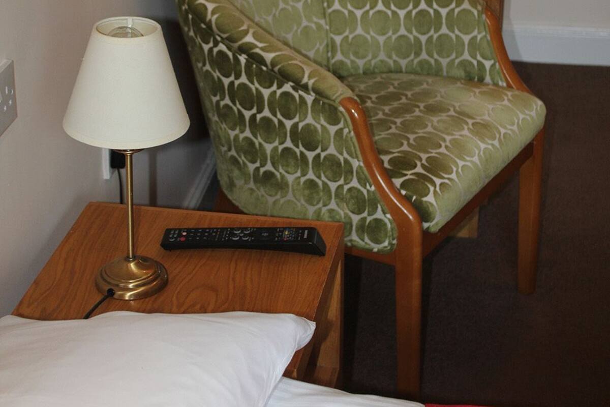 Ein Hotelzimmer mit einem Bett, einem Stuhl mit grünem gemusterten Kissen, einer Lampe und einer Fernbedienung auf einem Nachttisch aus Holz.