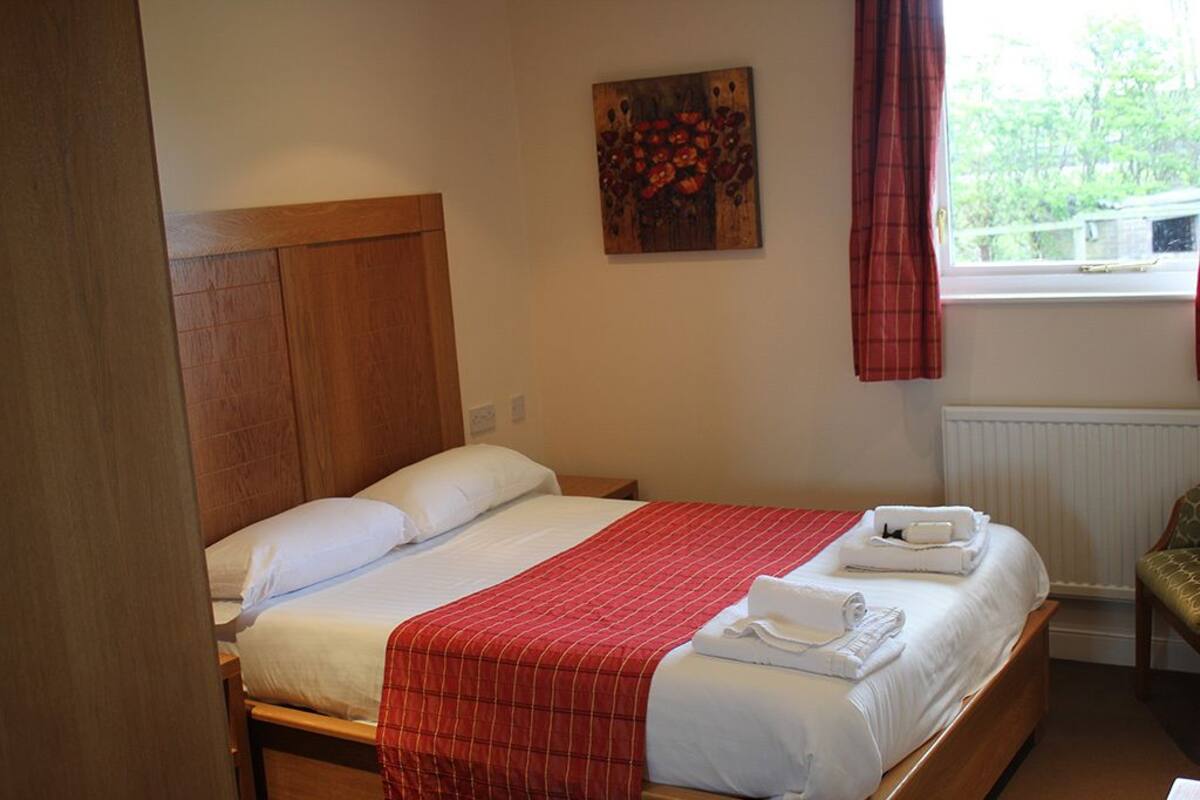 Ein ordentlich bezogenes Bett mit weißen Leinen und einer roten Karo-Decke, ein hölzernes Kopfende, ein Bild an der Wand und ein Fenster mit roten Vorhängen.