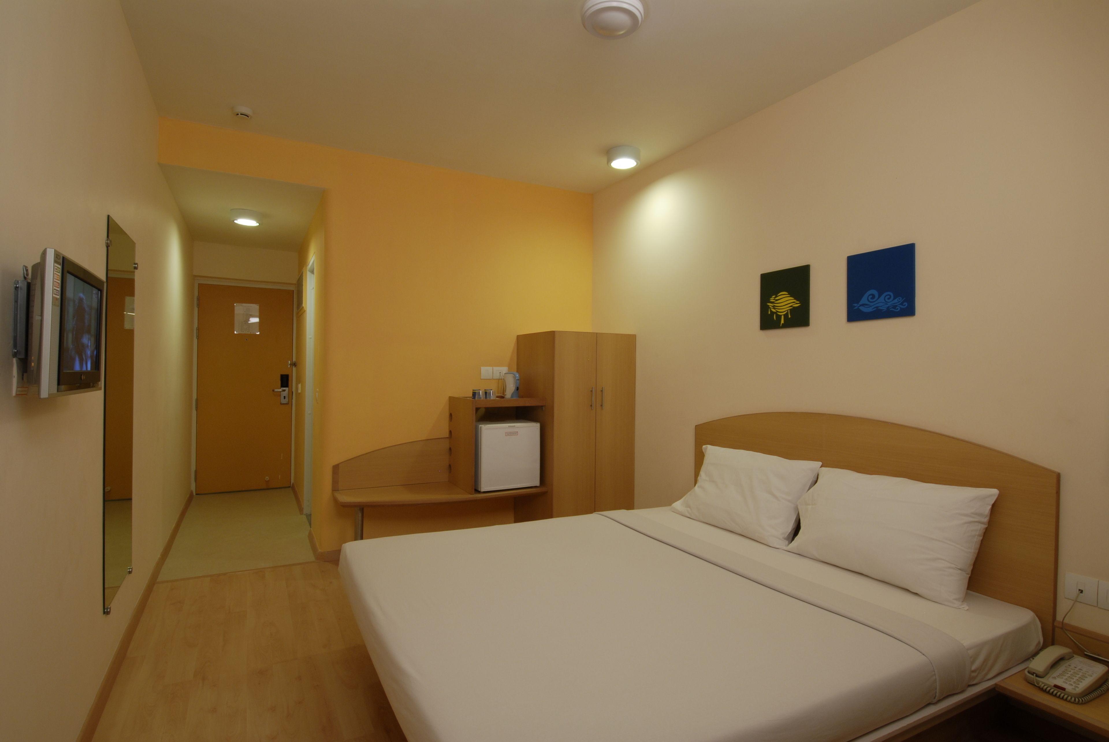 A hotel room with a large bed, a bedside table, a wardrobe, and a small desk with a lamp.