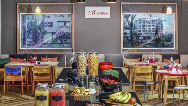Un'area buffet con diversi cibi e bevande, tavoli con sedie e due ampie finestre vista città.