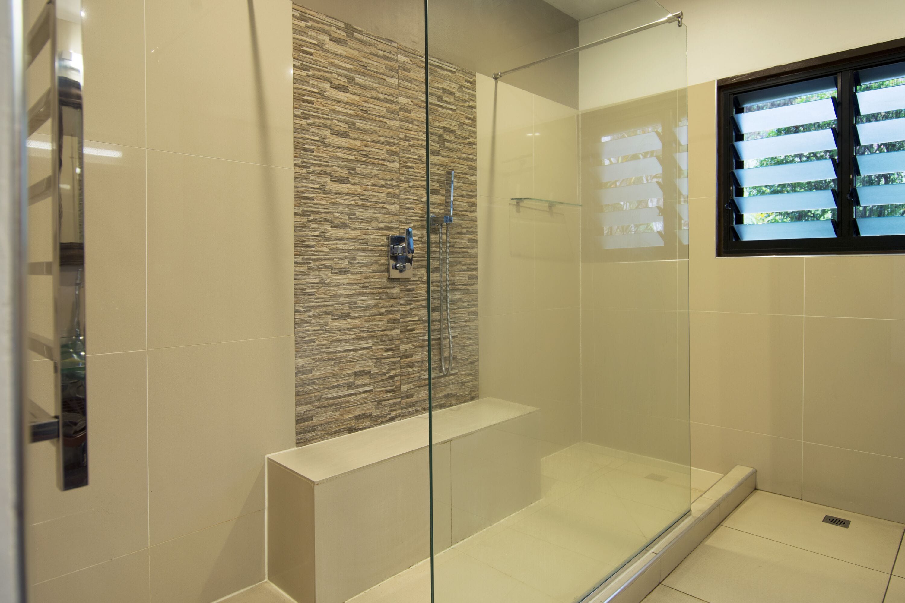 Un baño moderno con cabina de ducha de vidrio, una pared decorativa de piedra y una ventana con persianas.