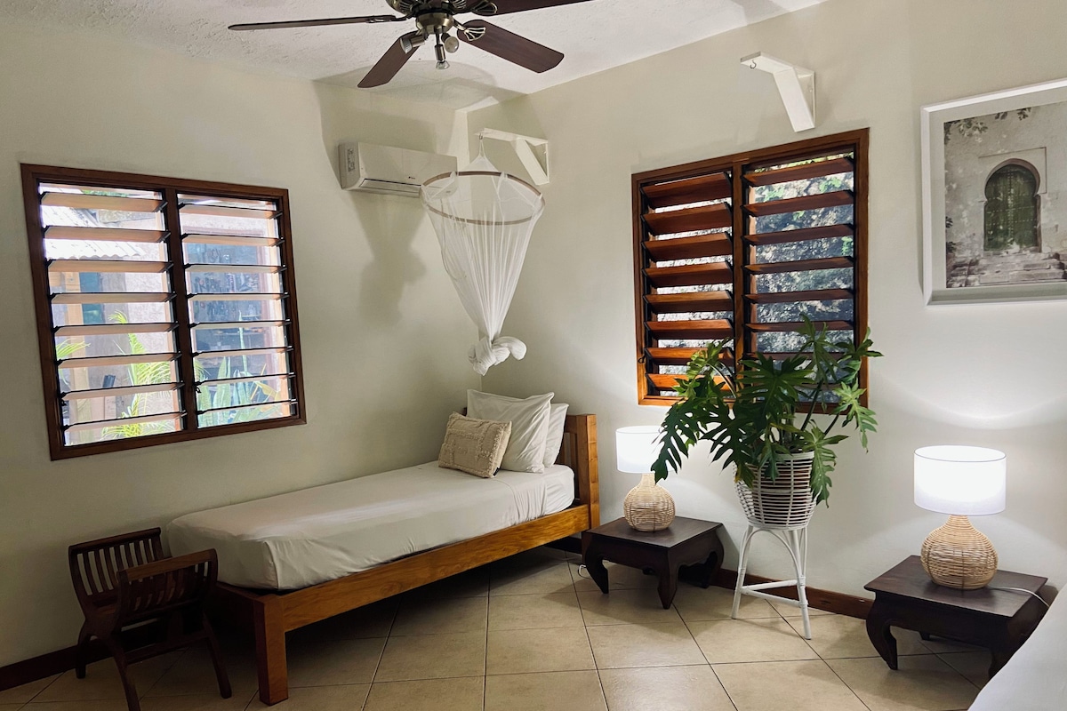Una habitación con una cama, ventilador de techo, ventana con persianas, planta en maceta y una lámpara.