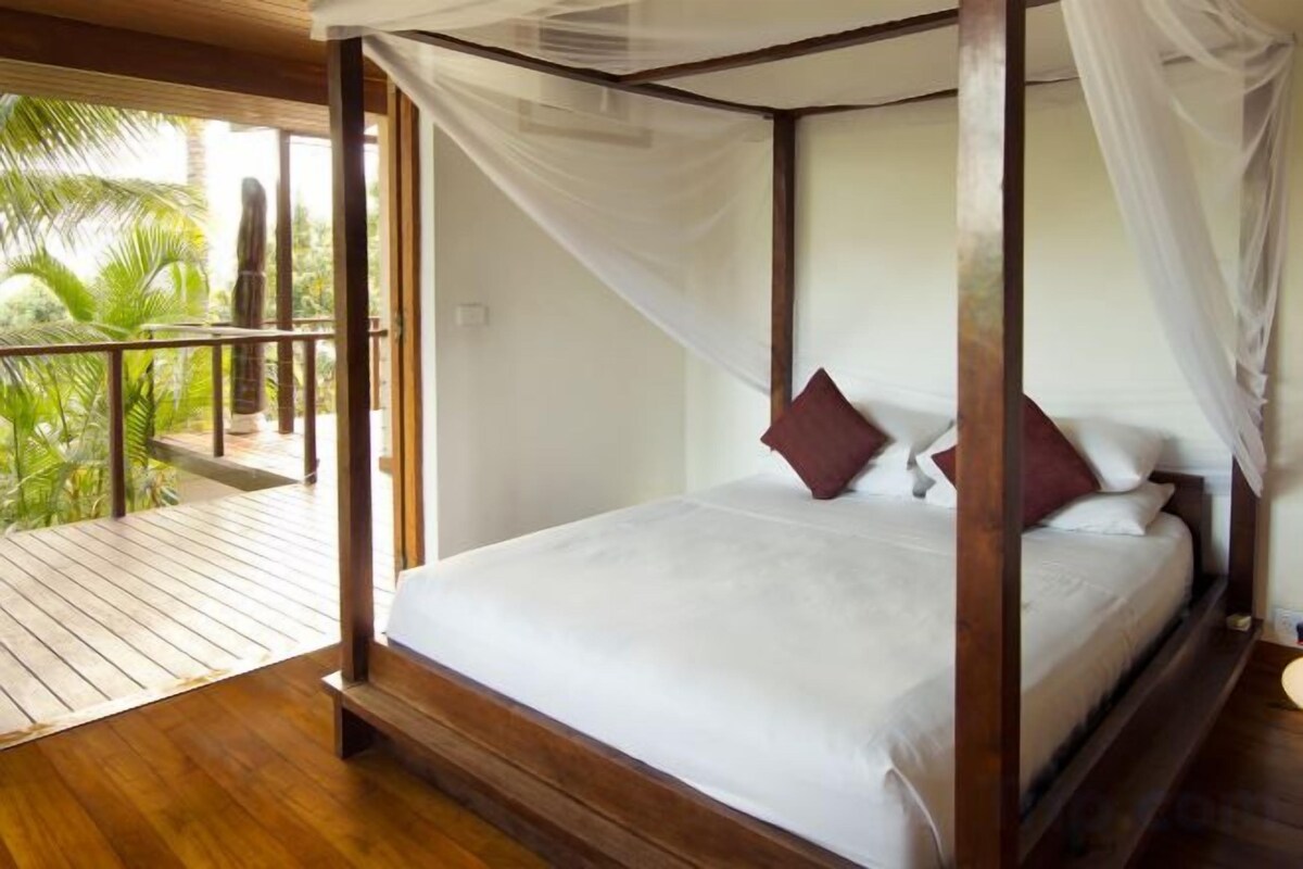 A four-poster bed with white bedding and red pillows, a wooden floor, and a balcony with a view of palm trees.