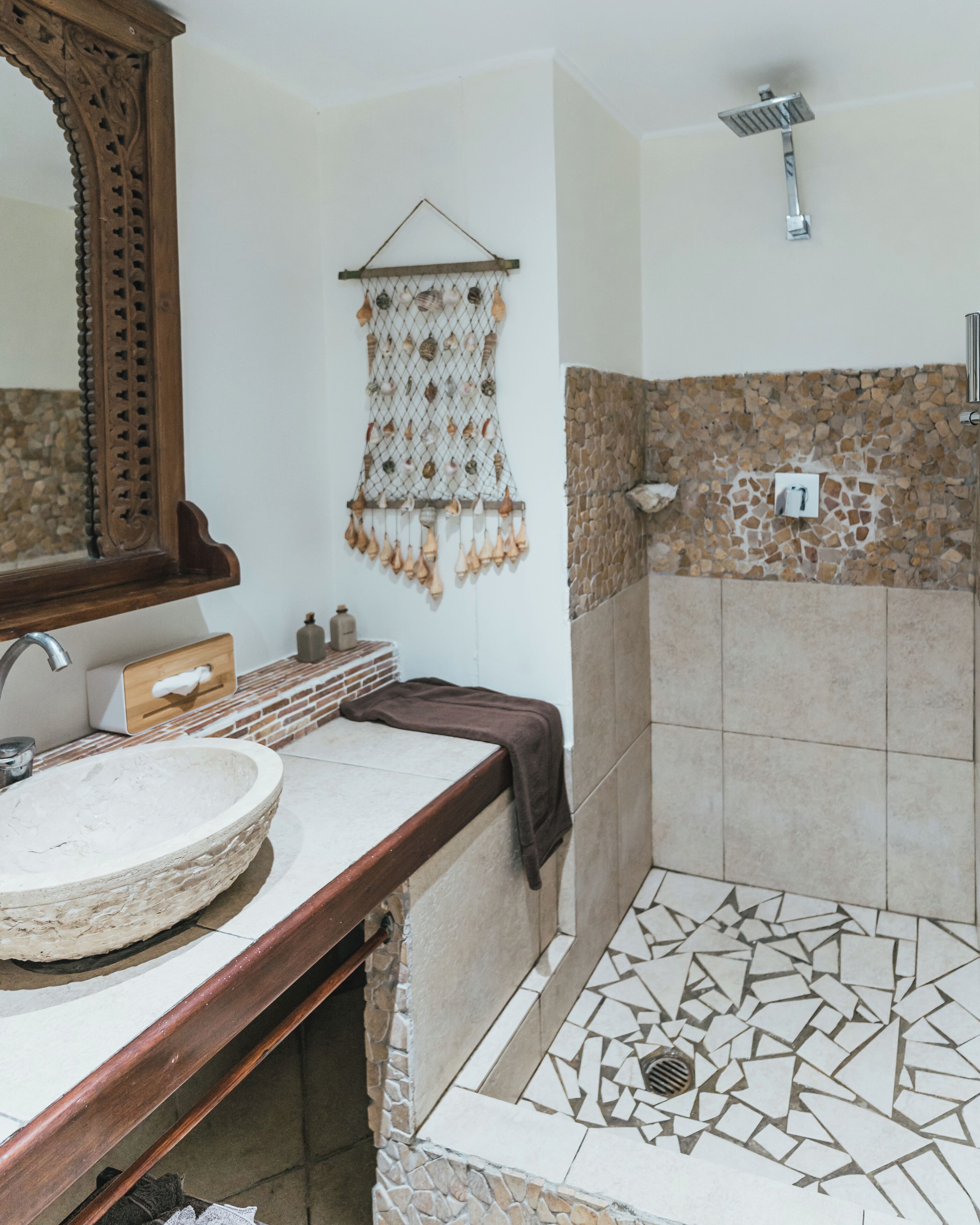 A bathroom with a round sink, a wooden vanity, a towel rack with decorative items, a shower with a stone wall, and a tiled floor.