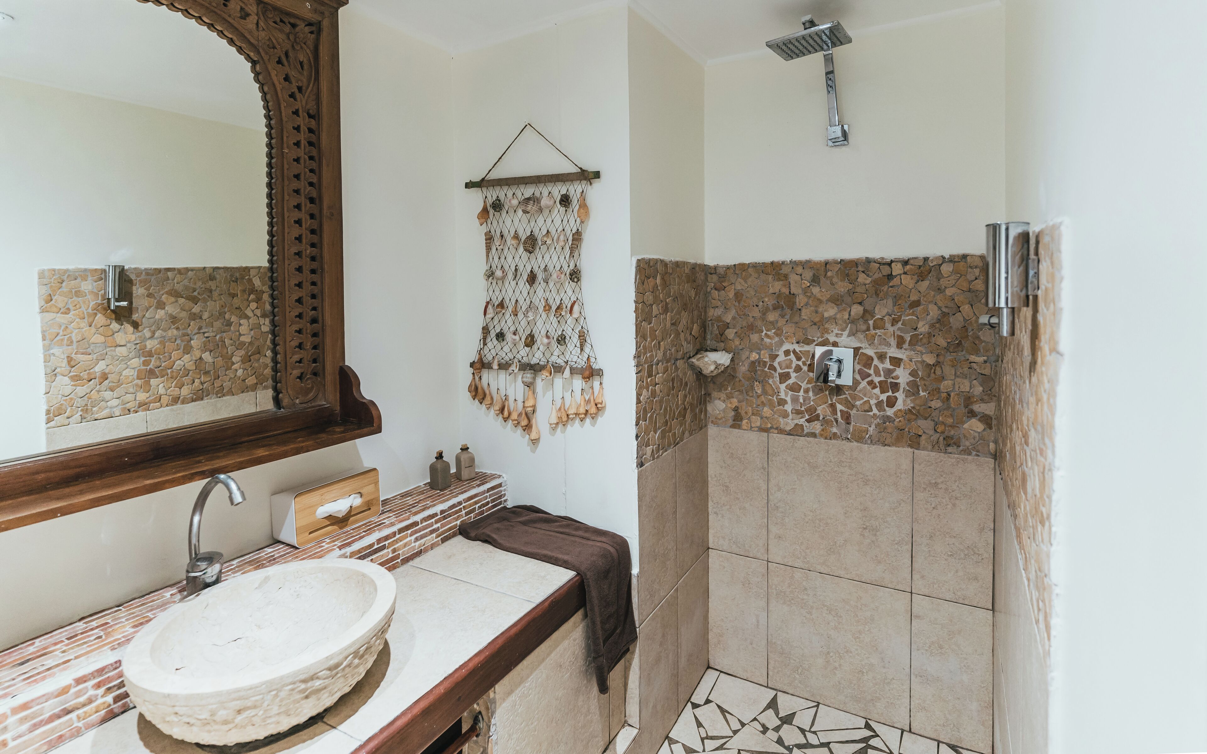 Un baño con pared de mosaico de piedra, tocador de madera, lavabo redondo, ducha con cabezal de lluvia y un adorno colgante.