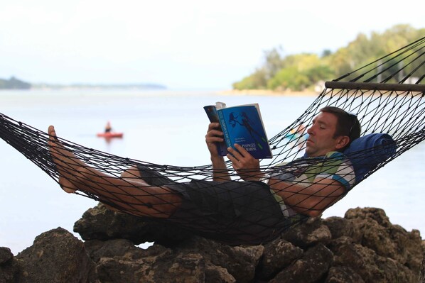 A person reclining in a hammock by a rocky shore with a body of water and a boat in the background.
