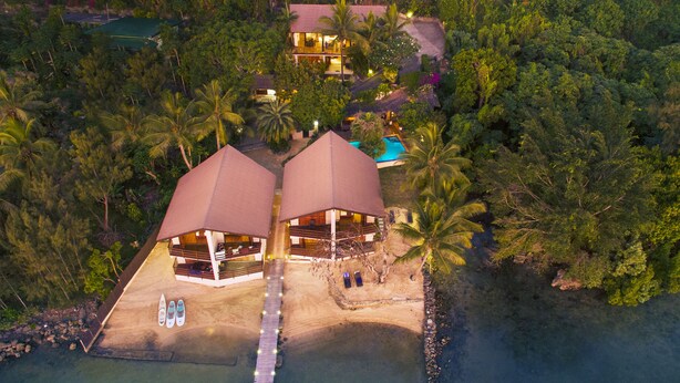 An aerial view of a resort with two large, covered structures, a dock, and a clear view of the surrounding tropical landscape.