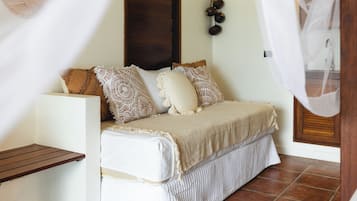 Una cama con cabecera blanca, varias almohadas y una colcha. A la izquierda hay un banco de madera y una decoración colgando sobre la cama.