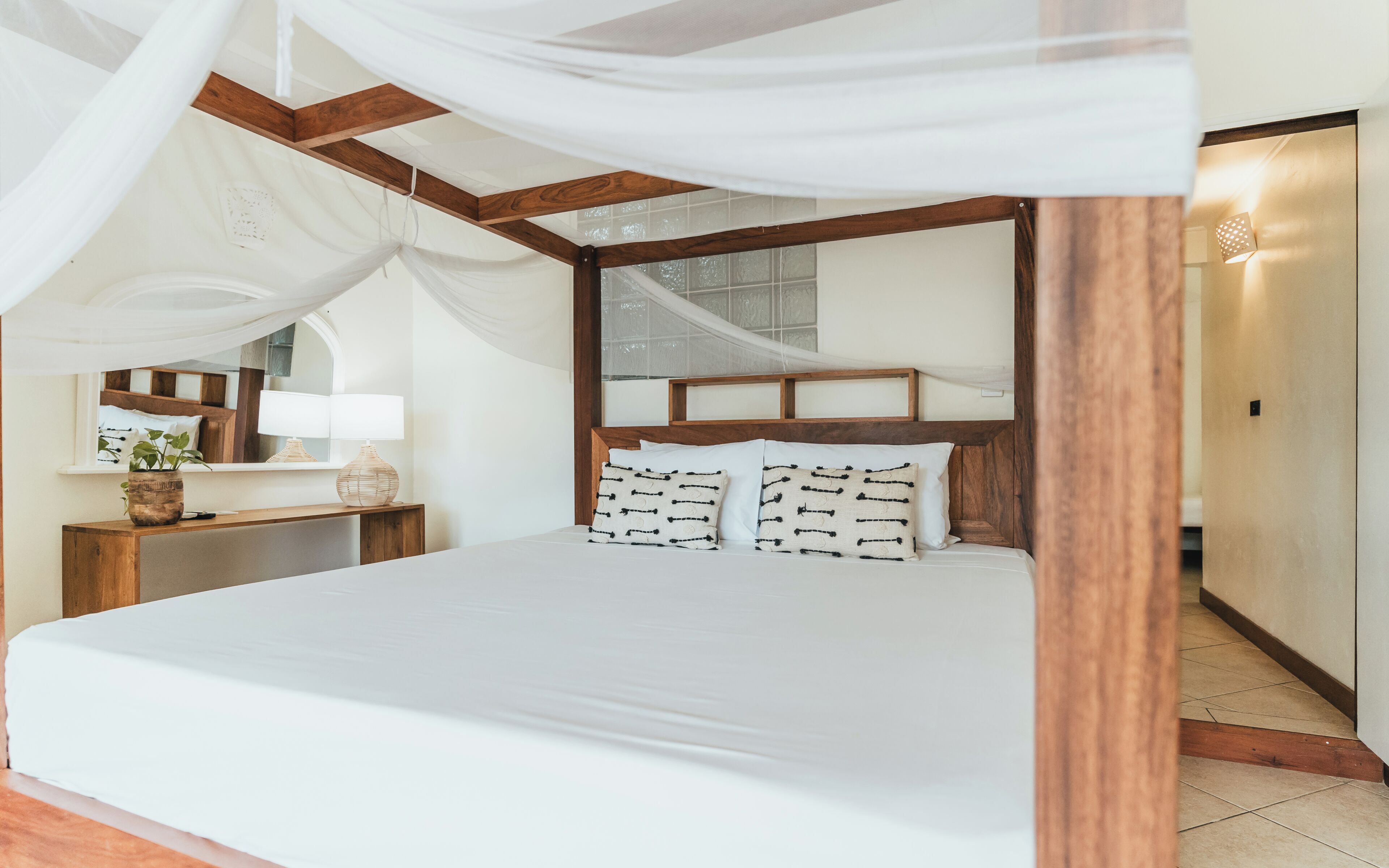 A bedroom with a canopy bed, wooden headboard, bedside table with lamp, and a view of a staircase.