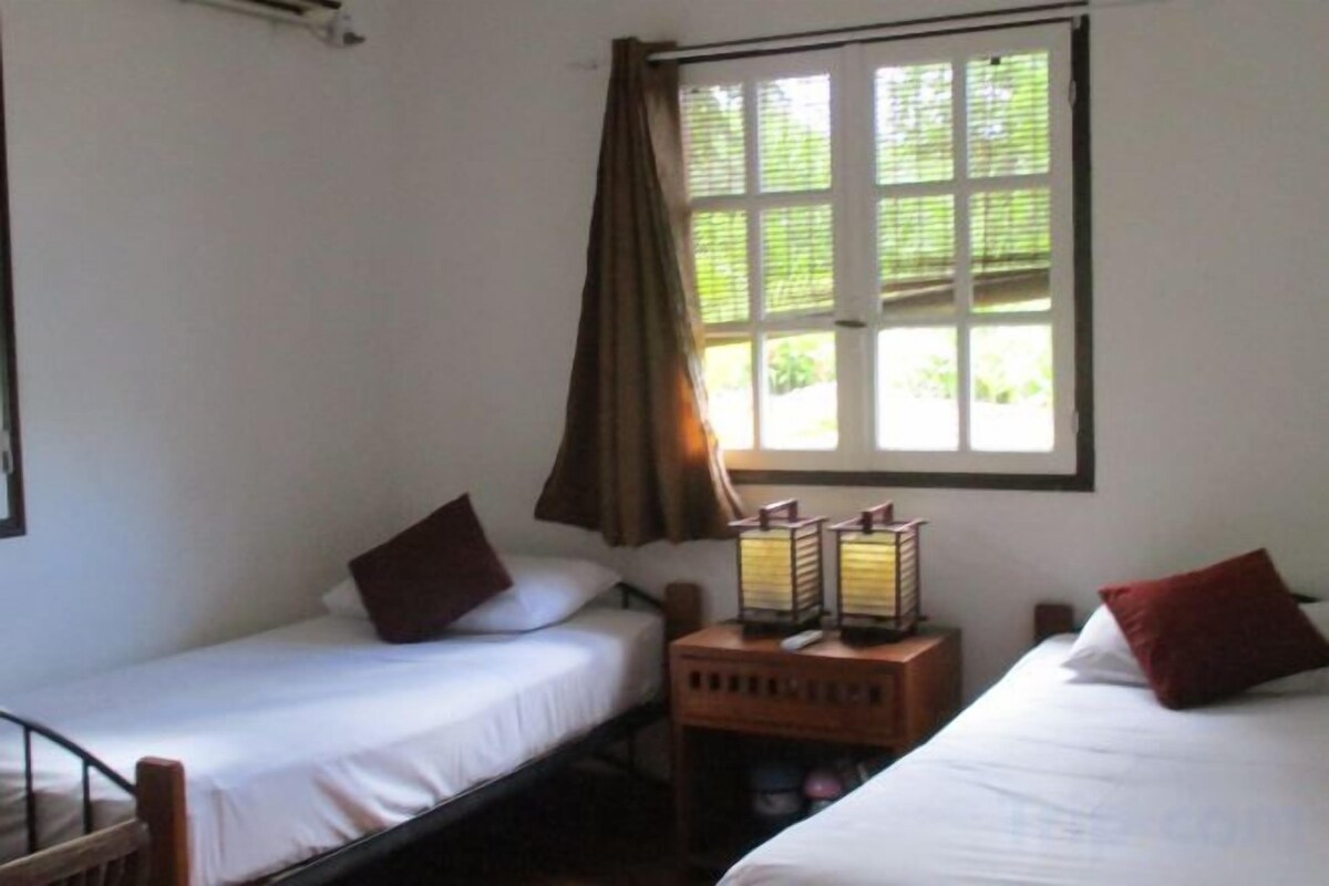 Una habitación con dos camas individuales, un ventanal con cortinas y una mesita de noche con lámpara.