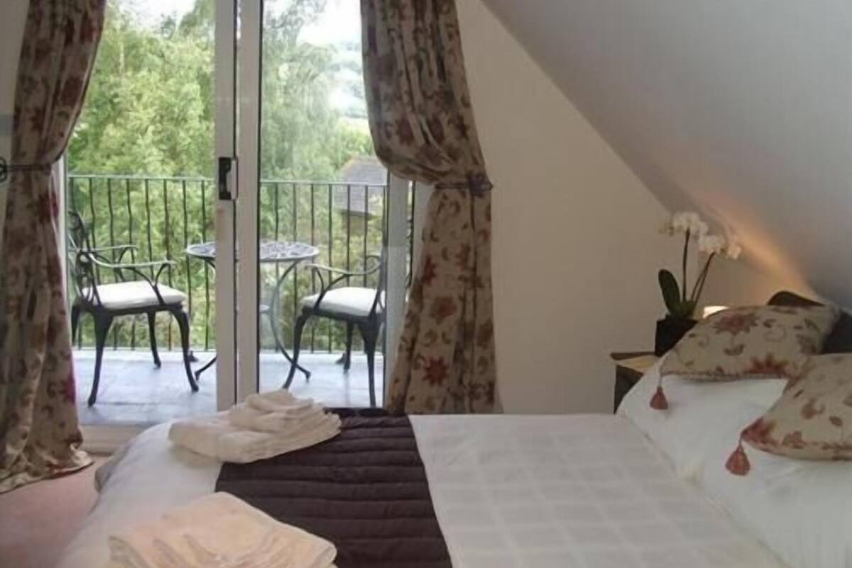 A bedroom with a bed, a nightstand, a potted plant, and a view of an outdoor balcony with chairs and a table.