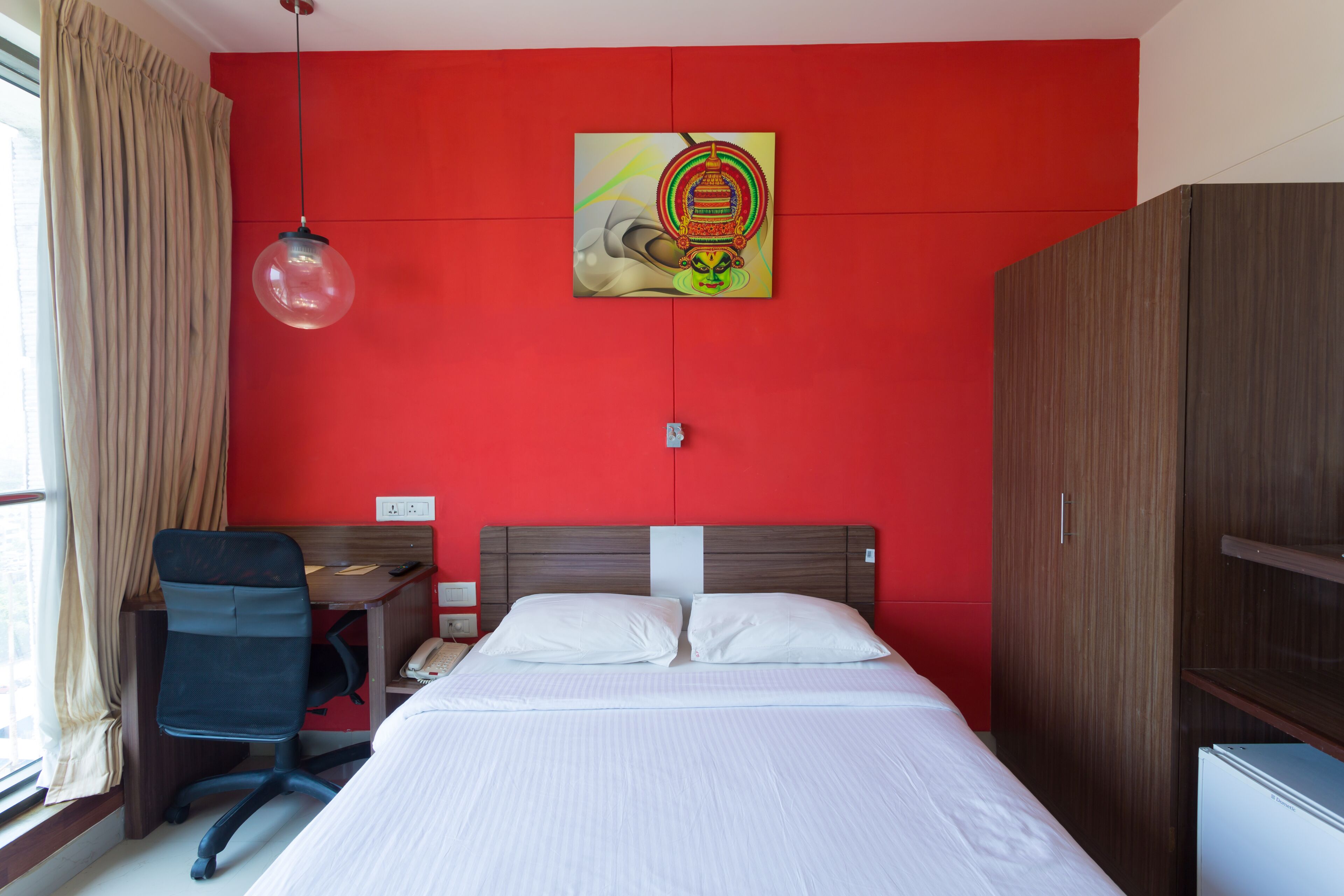 A hotel room with a red wall, a bed with white linens, a wooden wardrobe, a desk with a chair, and a hanging light fixture.