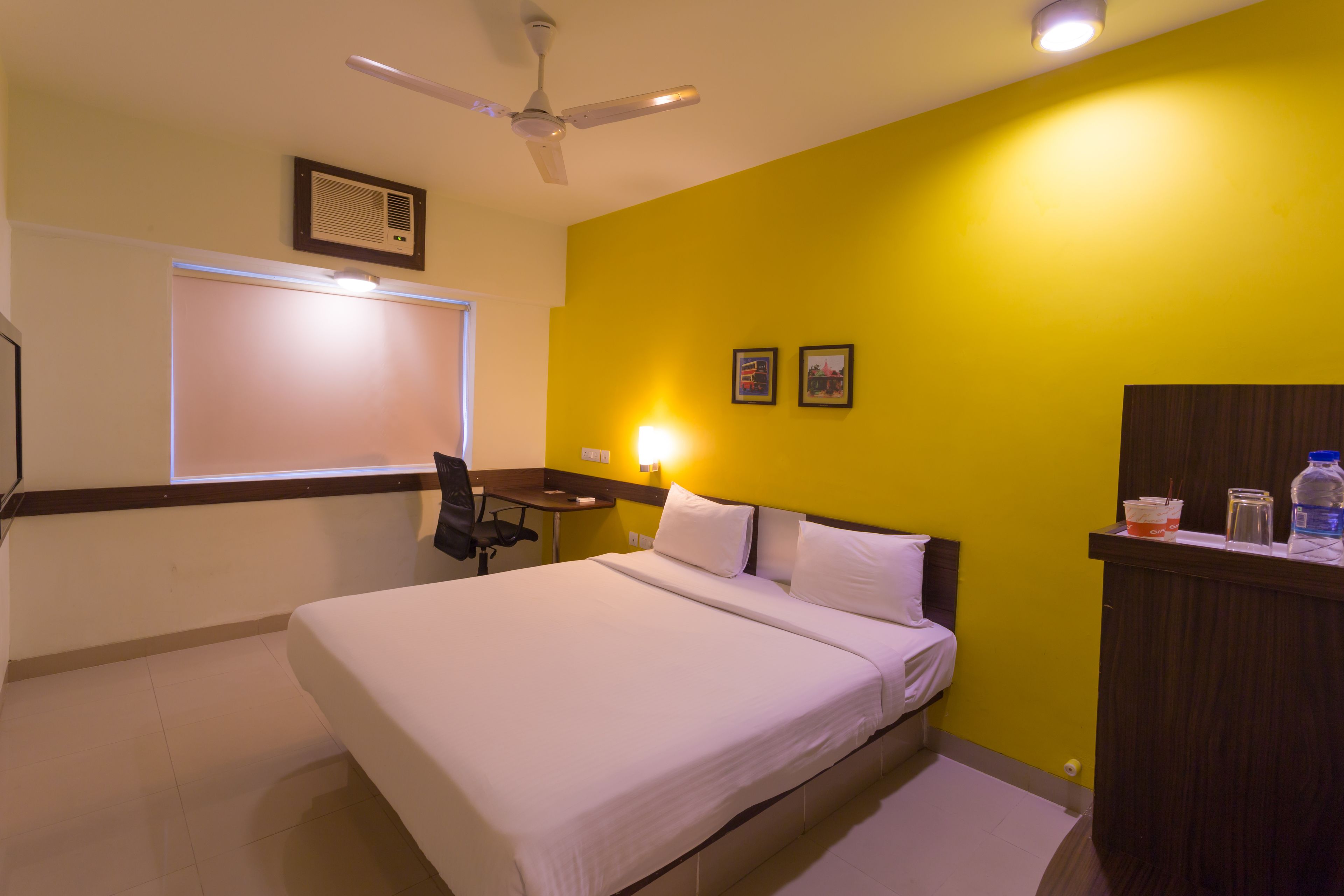 Habitación de hotel con cama, escritorio, silla, ventilador de techo, ventana y aire acondicionado instalado en la pared.