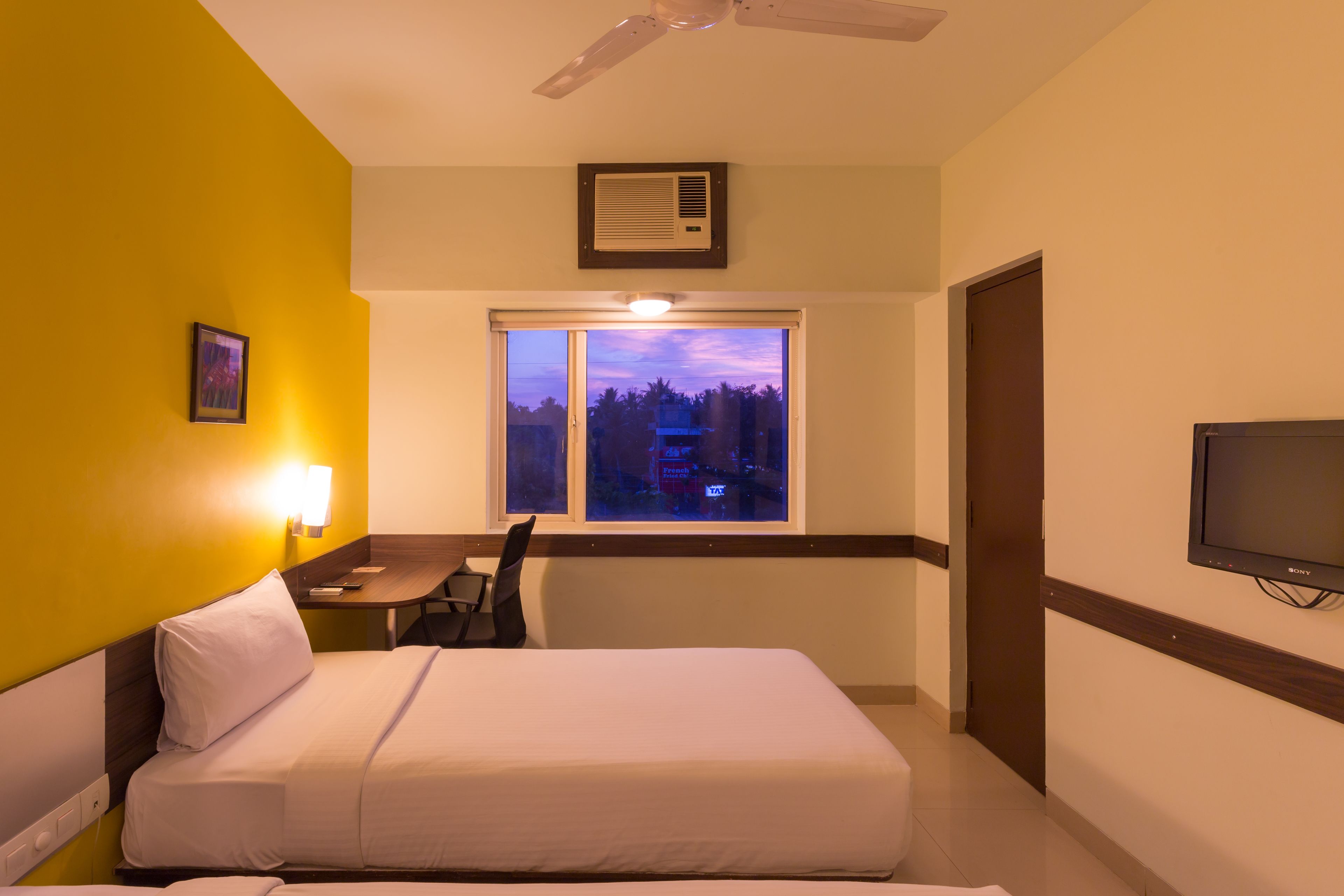 A hotel room with a bed, a desk, a chair, a TV, a window with a view, and an air conditioning unit.