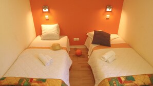 A hotel room with two single beds, each with a pillow, a rolled towel, and a hat on one bed. The room has an orange wall and a wooden floor. - VVF Sud Aveyron, Brusque (Brusque)