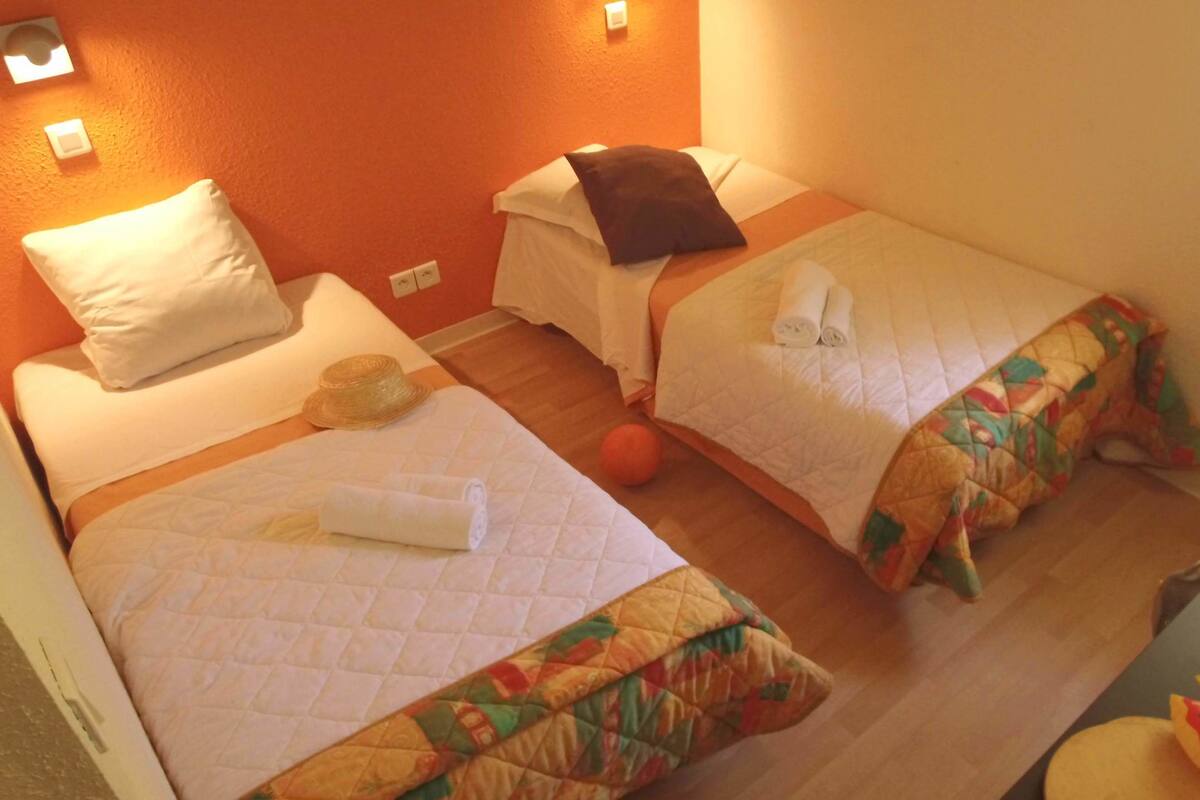 Een hotelkamer met twee bedden, elk voorzien van een gewatteerde sprei en kussens. Op de vloer tussen de bedden ligt een klein, rond, oranje object.